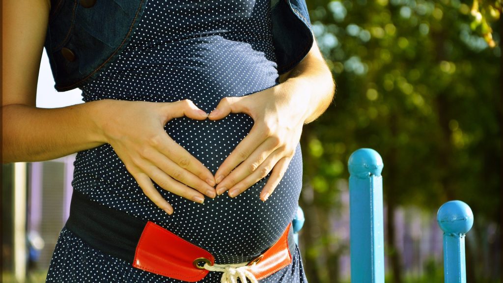 pregnant woman with a park and blue posts behind her, holding her hands in a heart shape over her pregnancy bump