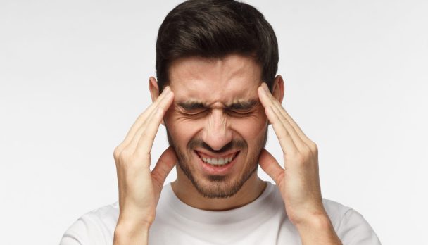 Portrait of young man isolated on grey background suffering from severe headache, pressing fingers to temples, closing eyes to relieve pain with helpless face expression