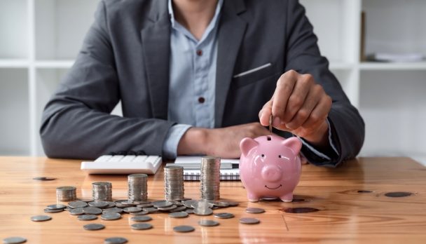 Businessman,Saving,Money,With,Piggy,Bank,And,Coins,On,Desk,